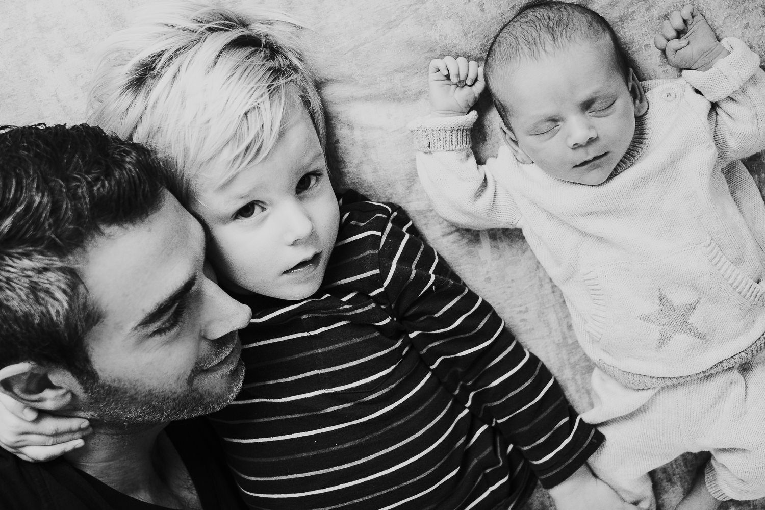 dad and big brother lying on bed with baby son black and white photo london newborn photography