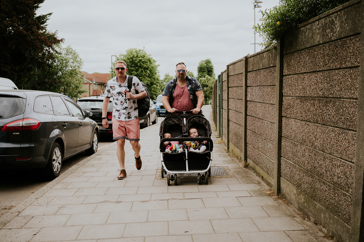 day in the life photo shoot two dads walking along pavement pushing a double buggy with two baby girls
