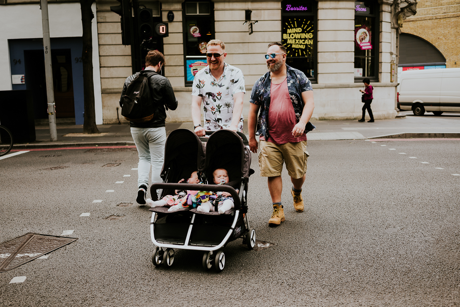 day in the life photo shoot two dads pushing double buggy with twin baby girls across pedestrian crossing smiling