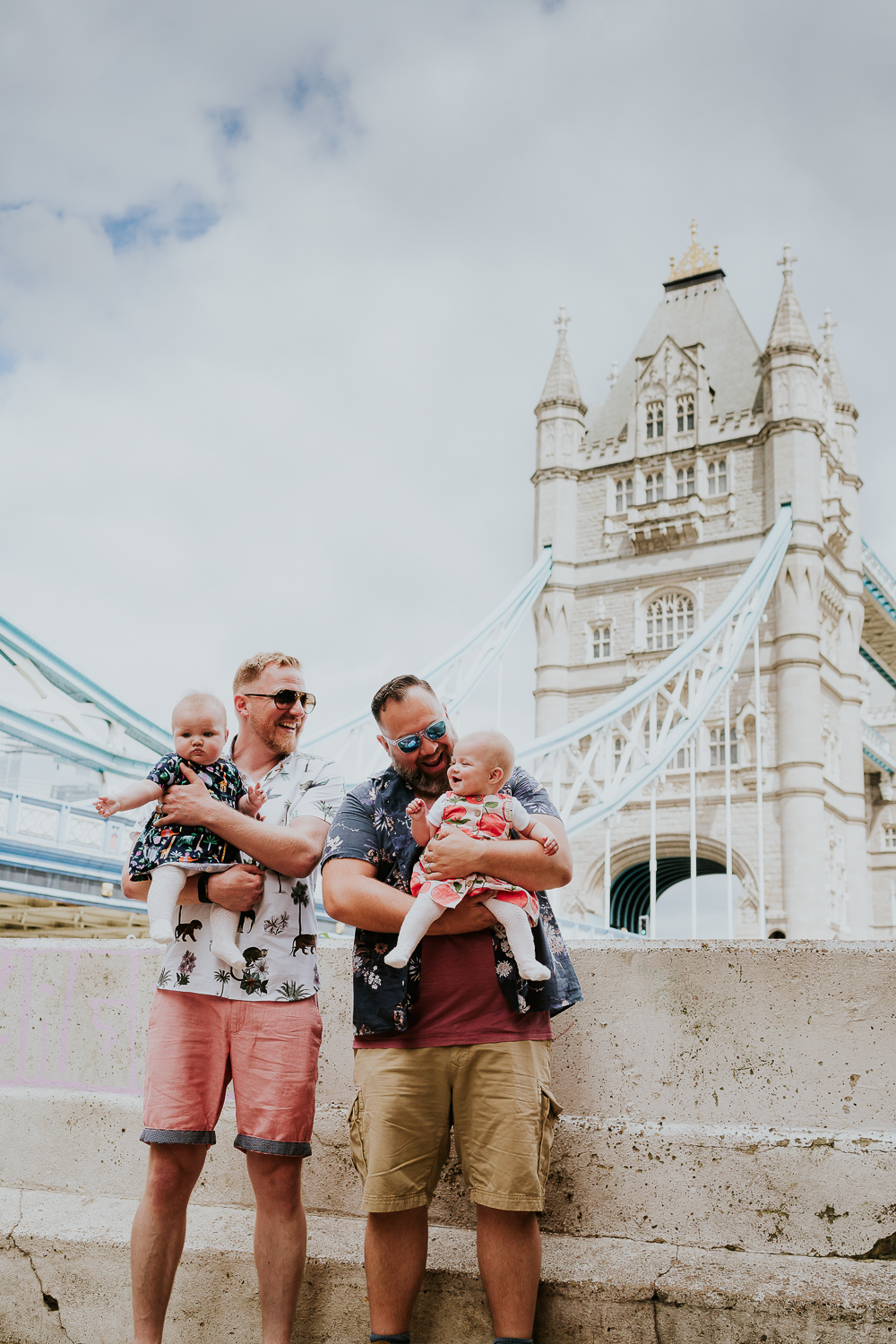 day in the life photo shoot two dads with twin baby girls standing with view of Tower Bridge in background