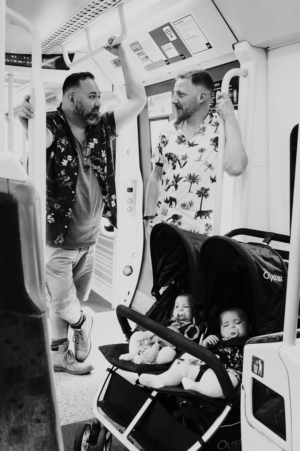 day in the life photo shoot two dads standing on train with double pushchair with twin baby girls