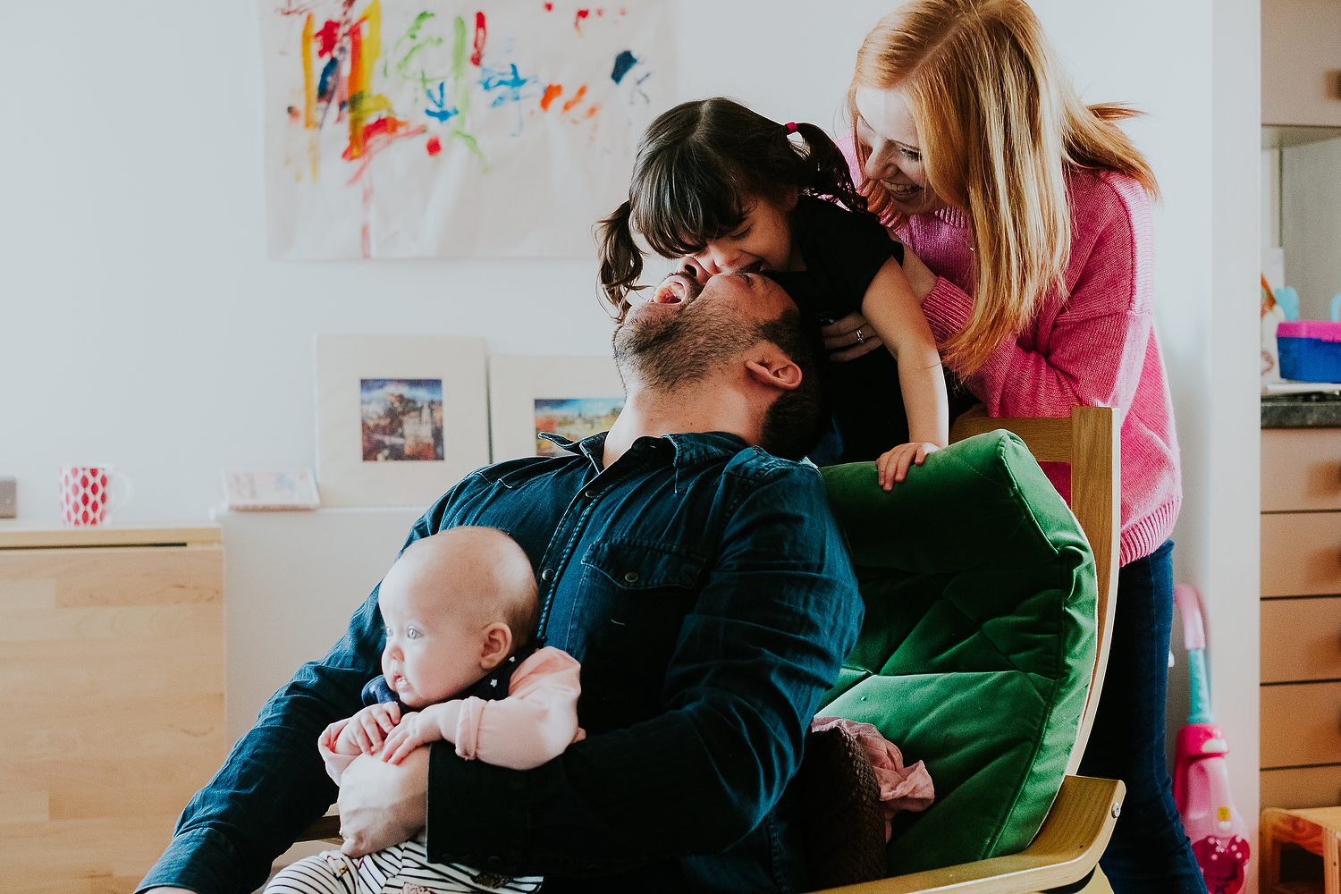 mother and father playing with baby girl and toddler daughter on chair at home islington family photographer