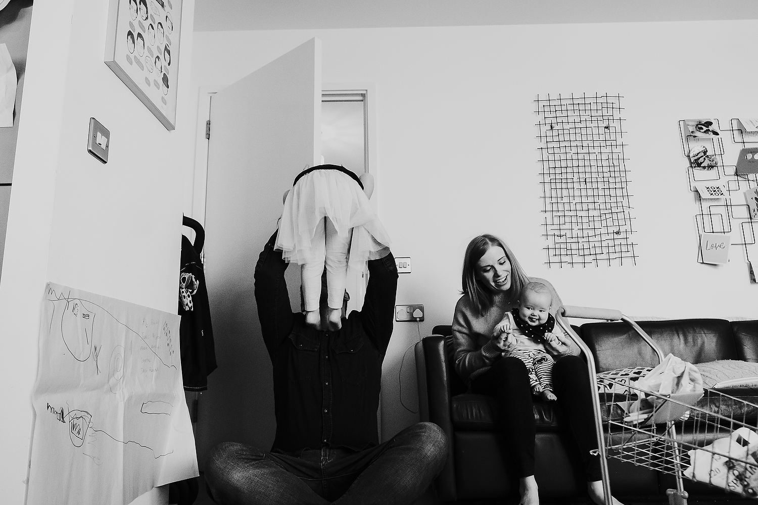 islington family photographer shot of dad holding daughter in the air and mother and baby sat on sofa playing at home