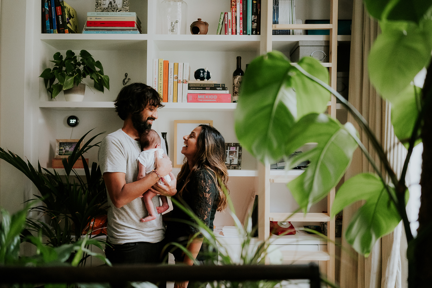 london newborn baby photographer parents holding baby daughter in home living space filled with bookshelves and plants