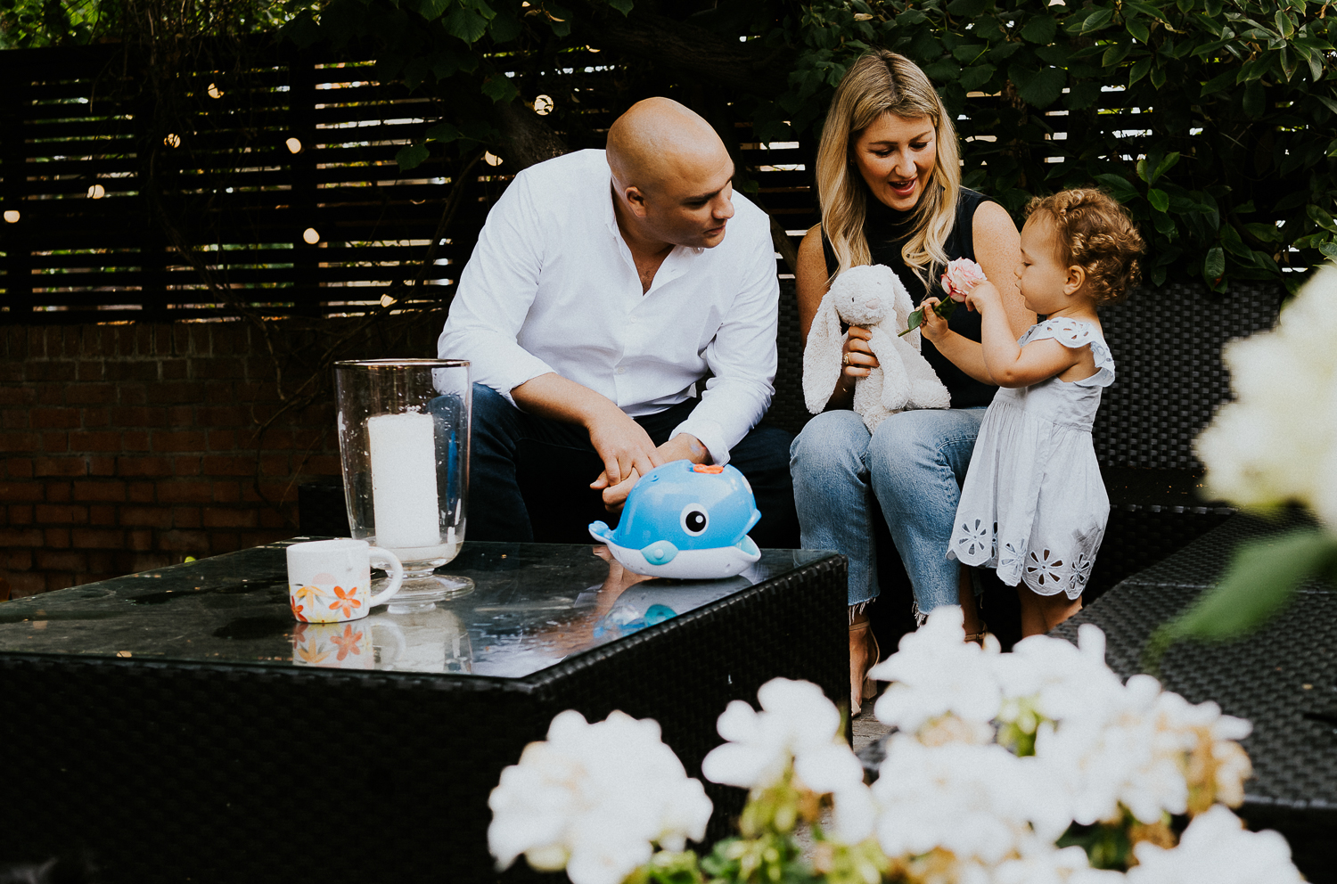 NORTH KENSINGTON FAMILY PHOTO SESSION parents and toddler daughter sitting on sofa in garden unposed portrait