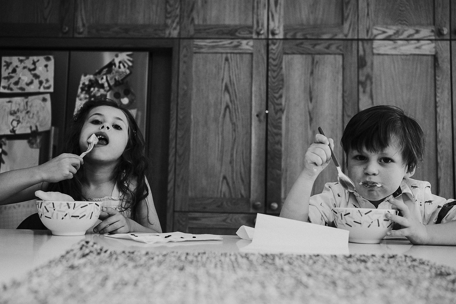 REAL LIFE FAMILY PHOTOS BLACK AND WHITE PHOTO OF BROTHER AND SISTER CHILDREN EATING DESSERT FROM BOWLS AT KITCHEN COUNTER