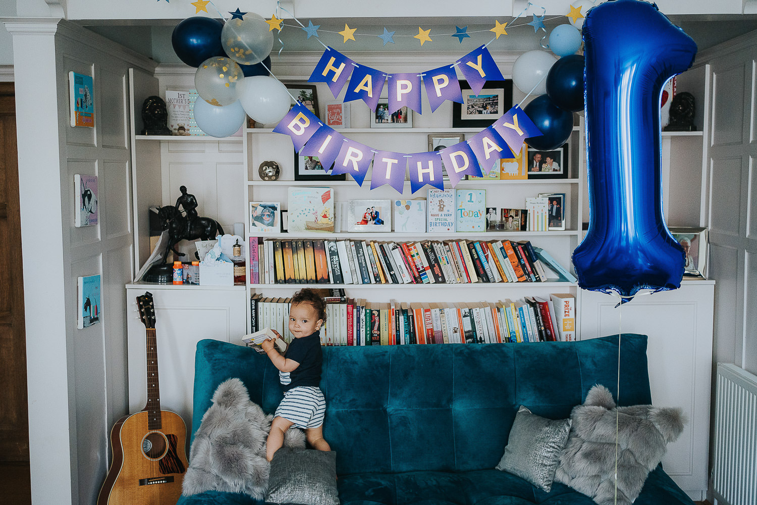 family photo shoot at home toddler boy stood on sofa with first birthday decorations around him and large bookshelf behind