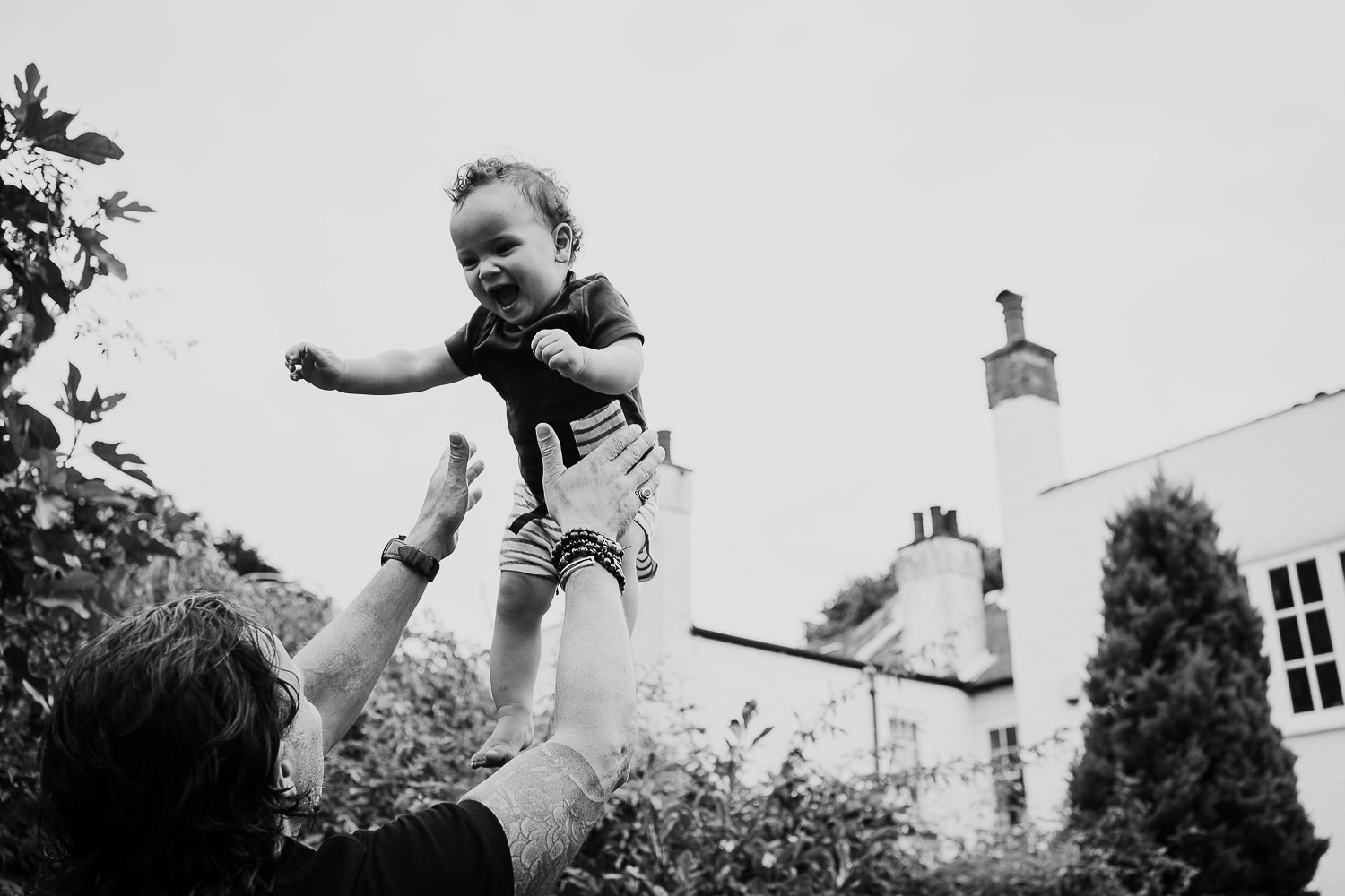 family photo shoot at home toddler boy being thrown in the air in the garden by his dad laughing