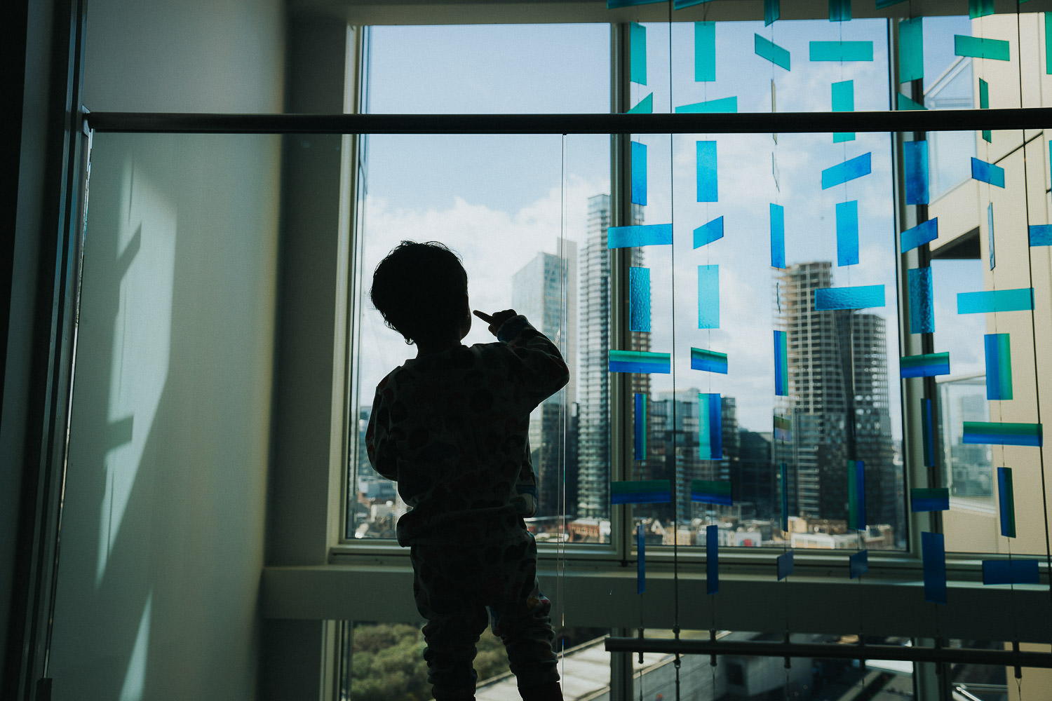 SHOREDITCH MATERNITY PHOTO SHOOT LITTLE TODDLER BOY LOOKING OUT OF LARGE APARTMENT BUILDING WINDOWS AT SKYSCRAPERS OUTSIDE