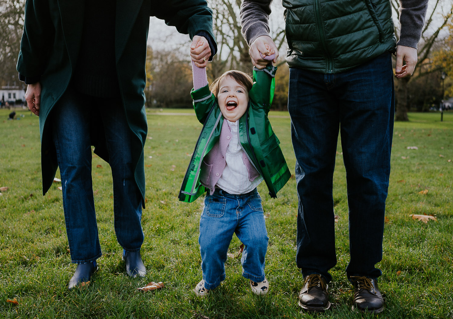 autumn mini photo shoot little toddler girl being swung between parents while walking in the park
